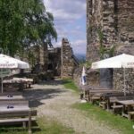 Sitzbereich im Freien mit Holztischen und -bänken unter weißen Sonnenschirmen, neben Steinruinen und einem Baum.