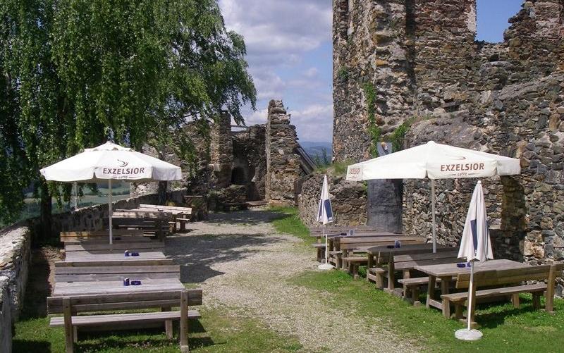 Sitzbereich im Freien mit Holztischen und -bänken unter weißen Sonnenschirmen, neben Steinruinen und einem Baum.