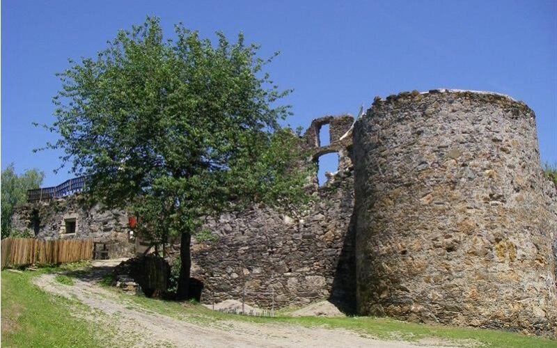 Die steinernen Ruinen eines alten Gebäudes mit einem zylindrischen Turm und einem Baum, der in der Nähe wächst, stehen unter einem klaren blauen Himmel.