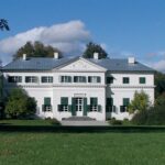 Ein weißes, zweistöckiges Haus mit grünen Fensterläden, einem zentralen Eingang und einem dreieckigen Giebel inmitten von Bäumen und einer großen Rasenfläche unter einem teilweise bewölkten Himmel.