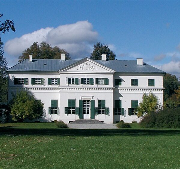 Ein weißes, zweistöckiges Haus mit grünen Fensterläden, einem zentralen Eingang und einem dreieckigen Giebel inmitten von Bäumen und einer großen Rasenfläche unter einem teilweise bewölkten Himmel.