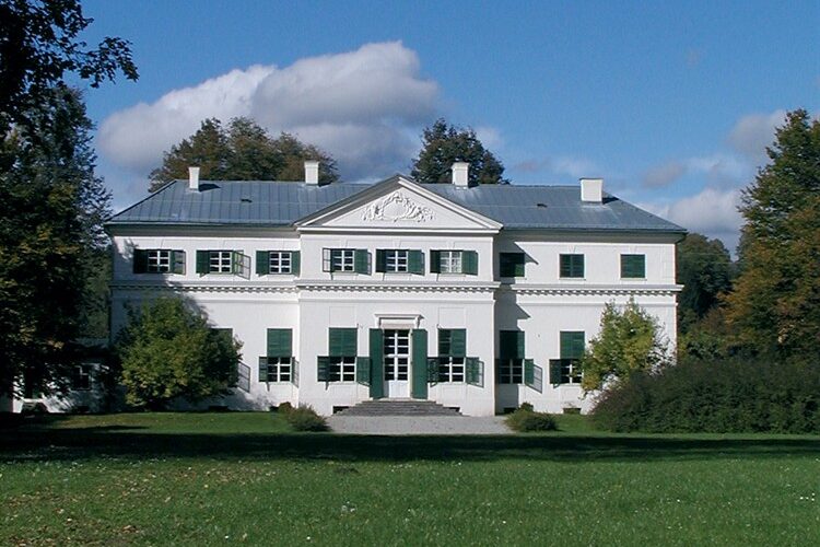 Ein weißes, zweistöckiges Haus mit grünen Fensterläden, einem zentralen Eingang und einem dreieckigen Giebel inmitten von Bäumen und einer großen Rasenfläche unter einem teilweise bewölkten Himmel.