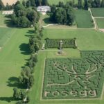 Luftaufnahme eines Heckenlabyrinths mit einem Hirsch und einem "rosegg"-Motiv, umgeben von grünen Feldern, Bäumen und einem großen Herrenhaus im Hintergrund.