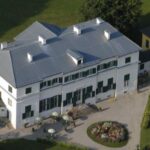 Luftaufnahme eines großen weißen zweistöckigen Gebäudes mit grünen Fensterläden, einem grauen Dach und einem kreisförmigen Blumengarten davor, umgeben von Gras und Bäumen.