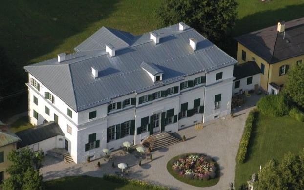 Luftaufnahme eines großen weißen zweistöckigen Gebäudes mit grünen Fensterläden, einem grauen Dach und einem kreisförmigen Blumengarten davor, umgeben von Gras und Bäumen.