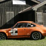 Ein orangefarbener Porsche-Rennwagen mit Sponsorenaufklebern steht auf dem Rasen vor einem dunklen Holzgebäude mit der Aufschrift PORSCHE" über der Tür.