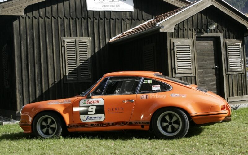Ein orangefarbener Porsche-Rennwagen mit Sponsorenaufklebern steht auf dem Rasen vor einem dunklen Holzgebäude mit der Aufschrift PORSCHE" über der Tür.