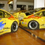 Gelber Rennwagen mit farbenfrohen Aufklebern, der in einem Innenraum geparkt ist und in großen Wandspiegeln reflektiert wird, die beide Seiten des Fahrzeugs zeigen.