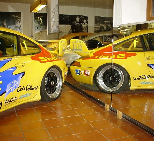 Gelber Rennwagen mit farbenfrohen Aufklebern, der in einem Innenraum geparkt ist und in großen Wandspiegeln reflektiert wird, die beide Seiten des Fahrzeugs zeigen.