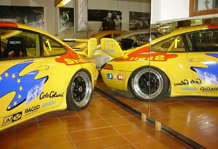 Gelber Rennwagen mit farbenfrohen Aufklebern, der in einem Innenraum geparkt ist und in großen Wandspiegeln reflektiert wird, die beide Seiten des Fahrzeugs zeigen.