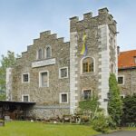 Steingebäude mit dekorativen weißen Akzenten, historischen Schildern, einem blau-gelben Emblem und einem Außenbereich mit alten Werkzeugen und Geräten auf der linken Seite.