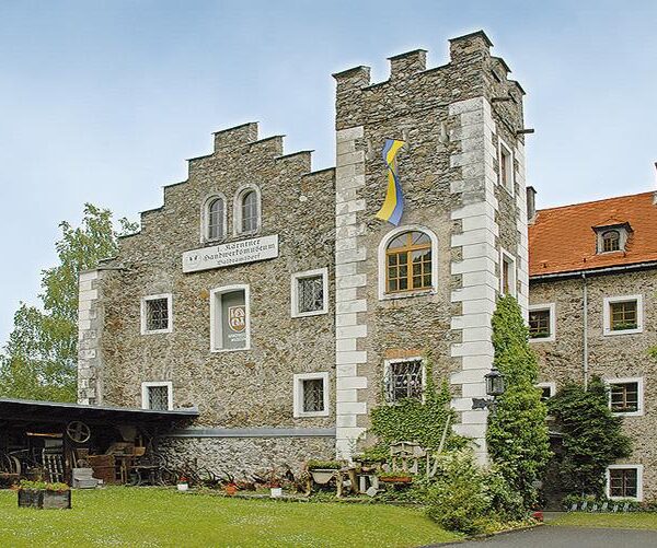 Steingebäude mit dekorativen weißen Akzenten, historischen Schildern, einem blau-gelben Emblem und einem Außenbereich mit alten Werkzeugen und Geräten auf der linken Seite.