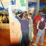 Mehrere Kinder versammeln sich um eine lehrreiche Ausstellung in einem Museum oder Naturzentrum mit Bildern von Bergen und Tieren an den Wänden und interagieren mit ihr.