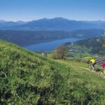 Zwei Personen fahren mit ihren Fahrrädern einen grasbewachsenen Hügel hinunter und überblicken einen See, ein Dorf und die Berge unter einem klaren blauen Himmel.