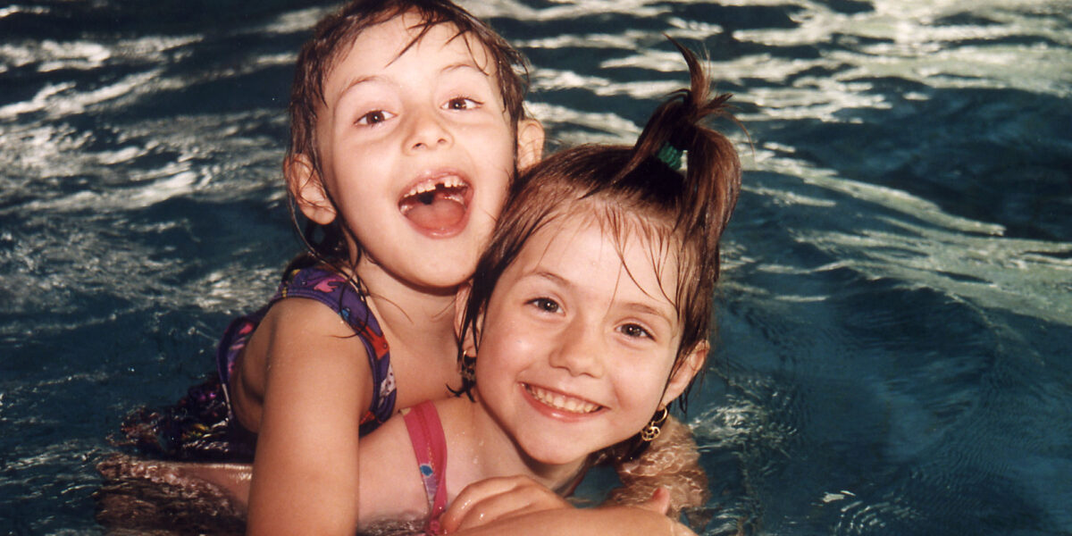 Zwei kleine Kinder in Badeanzügen lächeln und spielen beim Schwimmen in einem Pool, wobei ein Kind das andere von hinten umarmt.
