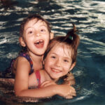 Zwei kleine Kinder in Badeanzügen lächeln und spielen beim Schwimmen in einem Pool, wobei ein Kind das andere von hinten umarmt.
