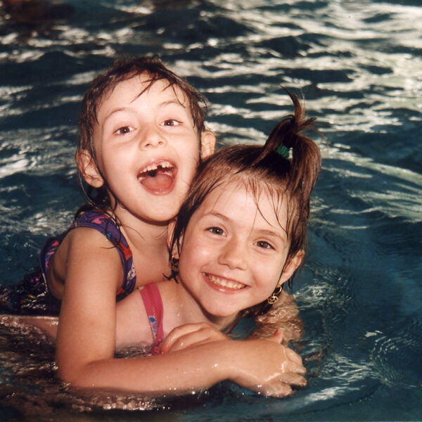 Zwei kleine Kinder in Badeanzügen lächeln und spielen beim Schwimmen in einem Pool, wobei ein Kind das andere von hinten umarmt.
