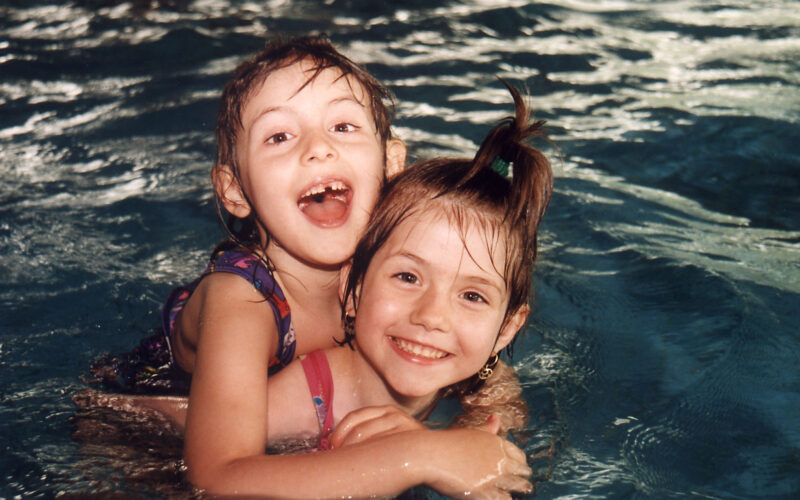 Zwei kleine Kinder in Badeanzügen lächeln und spielen beim Schwimmen in einem Pool, wobei ein Kind das andere von hinten umarmt.