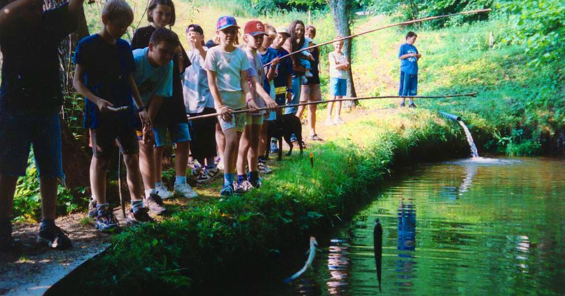 Eine Gruppe von Kindern steht mit Angelruten in der Hand an einem Teich, umgeben von grünen Bäumen und Gras. Ein Kind hat einen Fisch an Land gezogen.