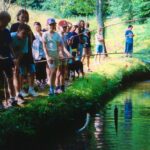 Eine Gruppe von Kindern steht mit Angelruten in der Hand an einem Teich, umgeben von grünen Bäumen und Gras. Ein Kind hat einen Fisch an Land gezogen.
