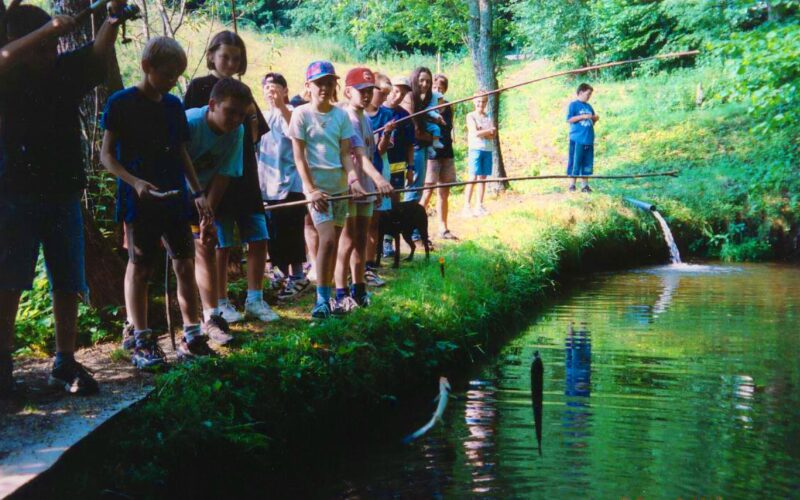 Eine Gruppe von Kindern steht mit Angelruten in der Hand an einem Teich, umgeben von grünen Bäumen und Gras. Ein Kind hat einen Fisch an Land gezogen.