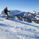 Ein Skifahrer, der einen Helm und eine Skibrille trägt, fährt einen verschneiten Berghang hinunter. Im Hintergrund sind schneebedeckte Gipfel unter einem klaren blauen Himmel zu sehen.
