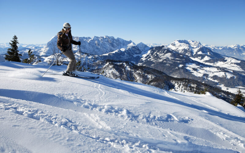Ein Skifahrer, der einen Helm und eine Skibrille trägt, fährt einen verschneiten Berghang hinunter. Im Hintergrund sind schneebedeckte Gipfel unter einem klaren blauen Himmel zu sehen.