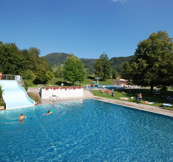 Freibad mit blauer Rutsche, umgeben von Gras, Bäumen und Hügeln; Menschen schwimmen und entspannen sich auf Handtüchern in der Sonne.