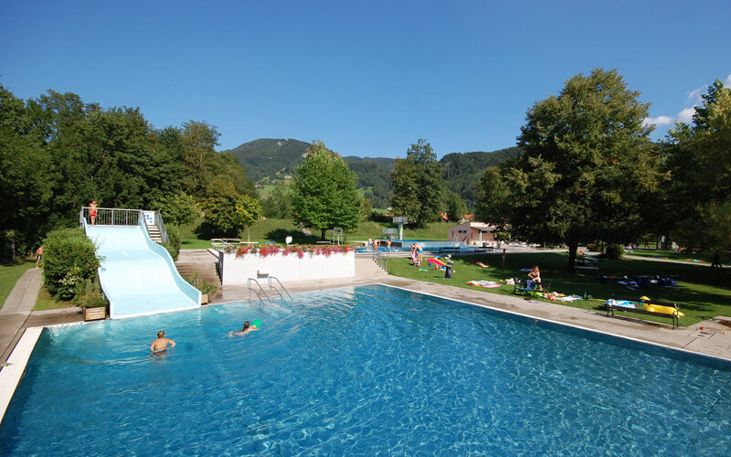 Freibad mit blauer Rutsche, umgeben von Gras, Bäumen und Hügeln; Menschen schwimmen und entspannen sich auf Handtüchern in der Sonne.