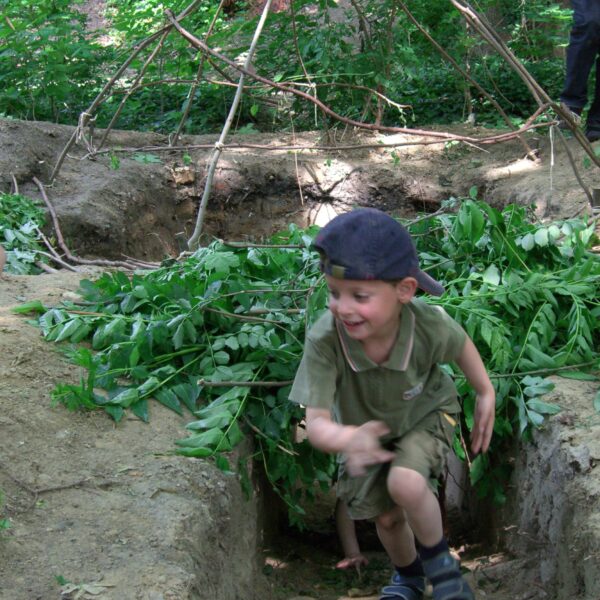 Ein kleiner Junge in grünem Outfit und marineblauer Mütze rennt durch einen schmutzigen Graben, der von belaubten Ästen umgeben ist, mit einer einfachen Holzkonstruktion im Hintergrund.