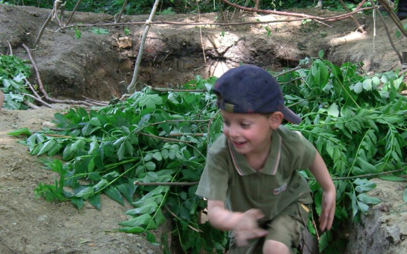 Ein kleiner Junge in grünem Outfit und marineblauer Mütze rennt durch einen schmutzigen Graben, der von belaubten Ästen umgeben ist, mit einer einfachen Holzkonstruktion im Hintergrund.