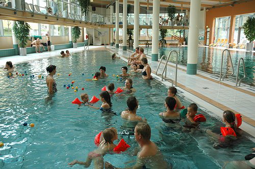Kinder und Erwachsene befinden sich in einem Hallenbad, einige mit Schwimmhilfen, während ein paar Leute am Beckenrand sitzen. Der Poolbereich ist geräumig, mit großen Fenstern und Pflanzen.