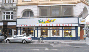 Straßenansicht eines Keramikmalereistudios mit dem Schild "Keramik selbst bemalen" und geparkten Autos davor.
