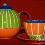 Ein farbenfrohes Teekannen- und Tassenset aus Keramik mit orangefarbenen, grünen, blauen und weißen Mustern vor einem roten Hintergrund.