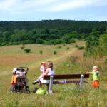 Eine Frau sitzt auf einer Bank in einer Wiese mit einem Baby auf dem Schoß, einem Kinderwagen in der Nähe und zwei kleinen Kindern, die in der Nähe stehen. Im Hintergrund sind bewaldete Hügel zu sehen.