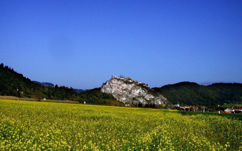 Ein felsiger Hügel mit einem Gebäude auf der Spitze erhebt sich über einem Feld mit gelben Blumen, mit bewaldeten Hügeln und einem klaren blauen Himmel im Hintergrund.