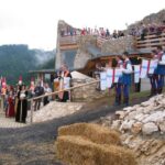 Menschen in mittelalterlichen Kostümen führen in einer steinernen Festung ein Reenactment auf, das von Holzplattformen und vom Boden aus beobachtet wird.