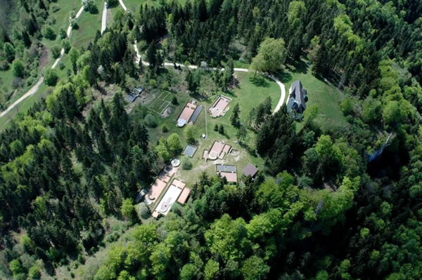 Luftaufnahme mehrerer Gebäude und einer Kapelle, umgeben von Bäumen und Grün, mit kurvenreichen Straßen, die die Landschaft durchziehen.