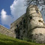 Ein verwitterter steinerner Burgturm mit kleinen Fenstern steht auf einem grasbewachsenen Hügel unter einem teilweise bewölkten Himmel.