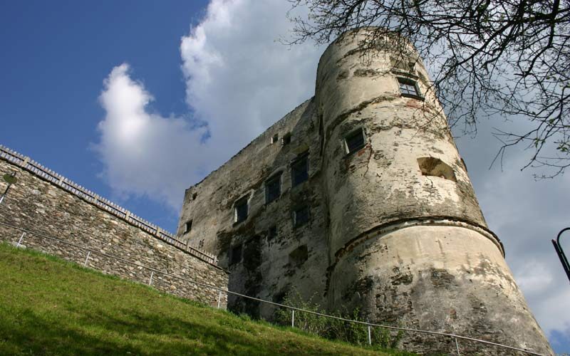 Ein verwitterter steinerner Burgturm mit kleinen Fenstern steht auf einem grasbewachsenen Hügel unter einem teilweise bewölkten Himmel.