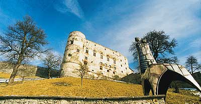 Ein steinernes Schloss auf einem grasbewachsenen Hügel ist aus einem niedrigen Winkel zu sehen, mit kahlen Bäumen und einer großen Metallskulptur im Vordergrund unter einem blauen Himmel.