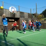 Eine Gruppe von Kindern spielt ein Ballspiel auf einem Sportplatz im Freien; ein Kind in Blau springt, um den Ball auf ein Tor zu werfen, während die anderen zusehen und reagieren.