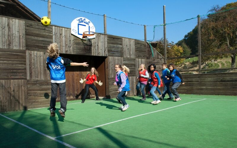 Eine Gruppe von Kindern spielt ein Ballspiel auf einem Sportplatz im Freien; ein Kind in Blau springt, um den Ball auf ein Tor zu werfen, während die anderen zusehen und reagieren.