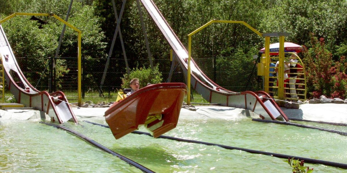 Eine Person fährt mit einem kleinen Boot eine Wasserrutsche hinunter, wobei das Boot knapp über dem Wasser in einem Vergnügungspark in der Luft schwebt.