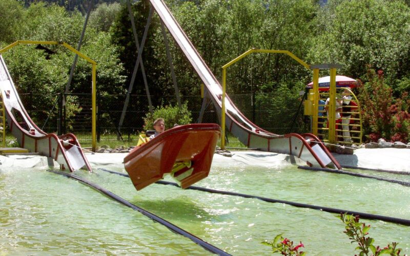 Eine Person fährt mit einem kleinen Boot eine Wasserrutsche hinunter, wobei das Boot knapp über dem Wasser in einem Vergnügungspark in der Luft schwebt.