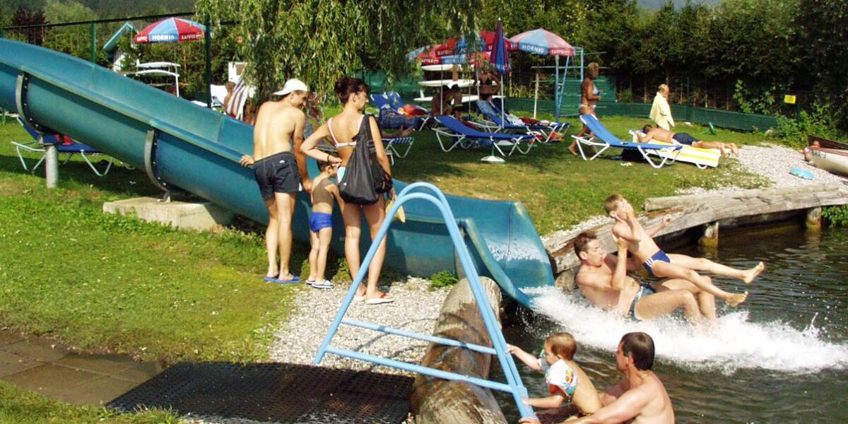 Mehrere Personen erfreuen sich an der Wasserrutsche und dem Pool im Freien, wo die Kinder im Wasser planschen und die Erwachsenen an einem sonnigen Tag in der Nähe Aufsicht führen.