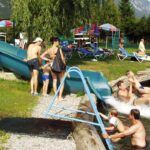 Mehrere Personen erfreuen sich an der Wasserrutsche und dem Pool im Freien, wo die Kinder im Wasser planschen und die Erwachsenen an einem sonnigen Tag in der Nähe Aufsicht führen.