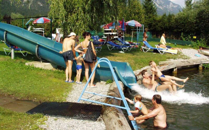Mehrere Personen erfreuen sich an der Wasserrutsche und dem Pool im Freien, wo die Kinder im Wasser planschen und die Erwachsenen an einem sonnigen Tag in der Nähe Aufsicht führen.