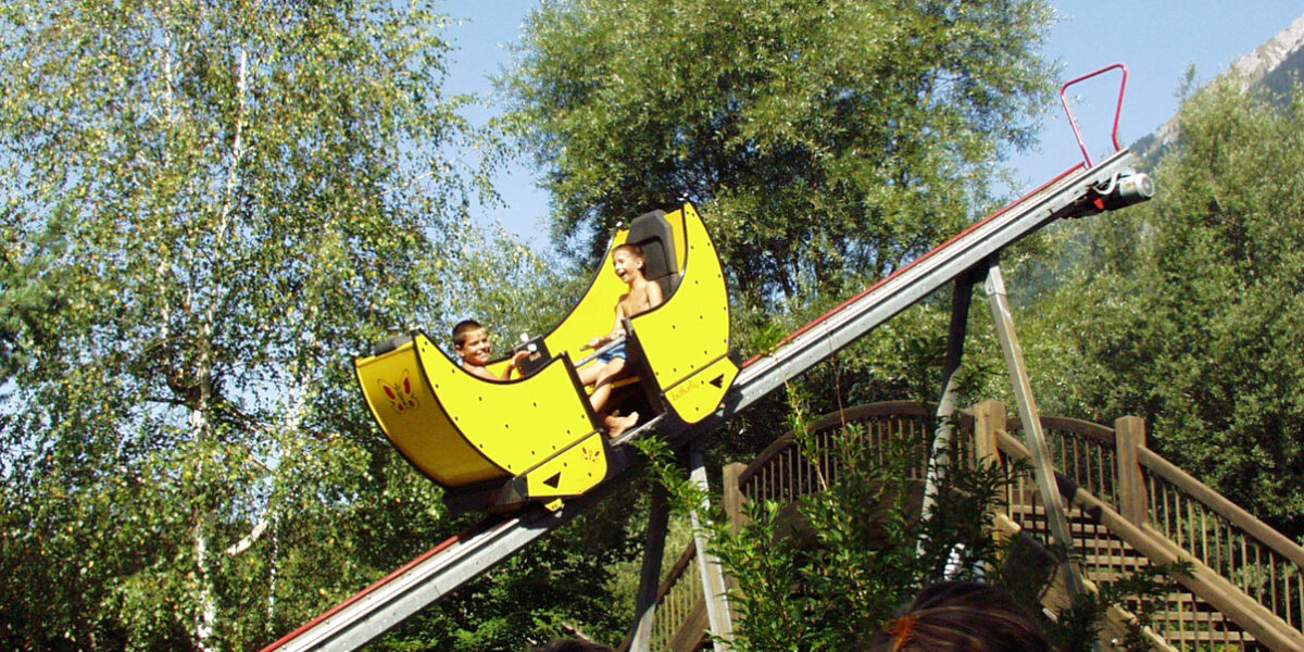 Zwei Personen fahren auf einer kleinen gelben Achterbahn eine abschüssige Strecke im Freien hinunter, wobei im Vordergrund Bäume und Zuschauer zu sehen sind.