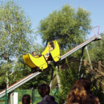 Zwei Personen fahren auf einer kleinen gelben Achterbahn eine abschüssige Strecke im Freien hinunter, wobei im Vordergrund Bäume und Zuschauer zu sehen sind.